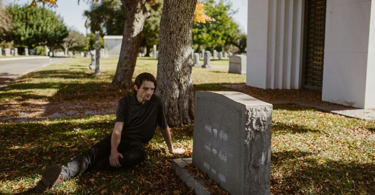 A man sits somberly beside a tombstone in a sunlit cemetery reflecting on loss and mourning