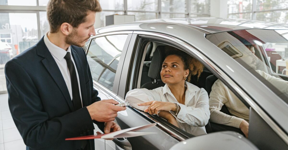 A car dealer hands keys to a woman sitting inside a vehicle in a car showroom