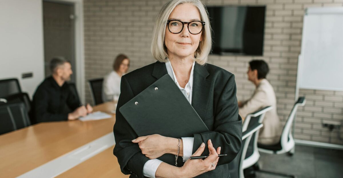 Senior businesswoman with eyeglasses holding a clipboard in a modern conference room with colleagues