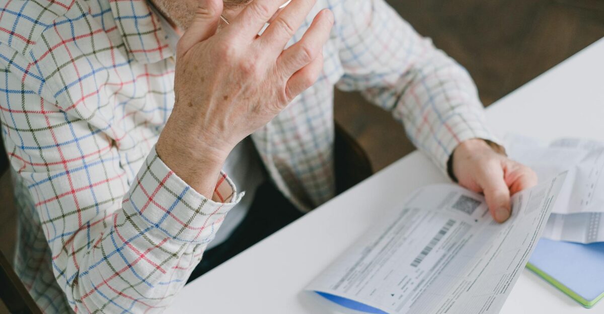 Elderly man in patterned shirt reading and holding bills at a home table appearing focused