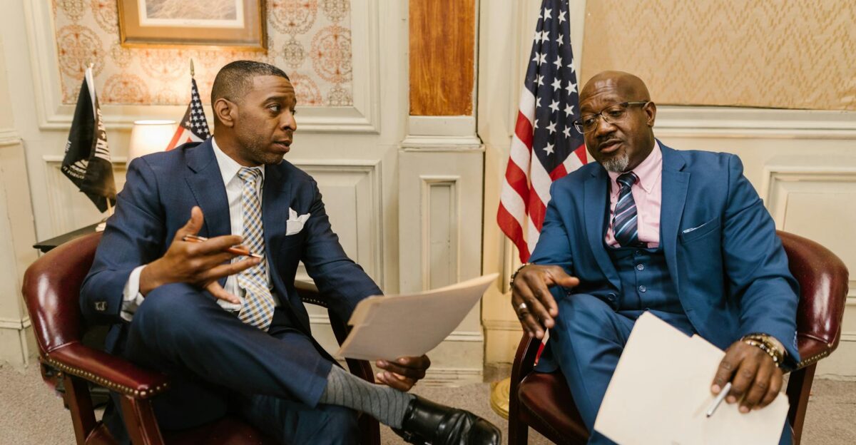 Two African American men in formal attire discussing documents in an office setting with flags in the background