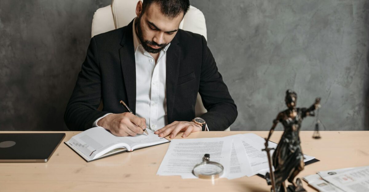Focused lawyer in black suit at desk writing on documents in an office setting with legal statue
