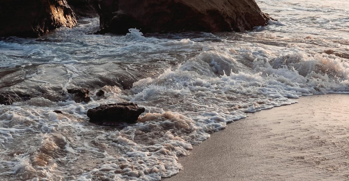 Serene view of waves striking rocks on a sandy beach during sunset capturing the ocean s beauty