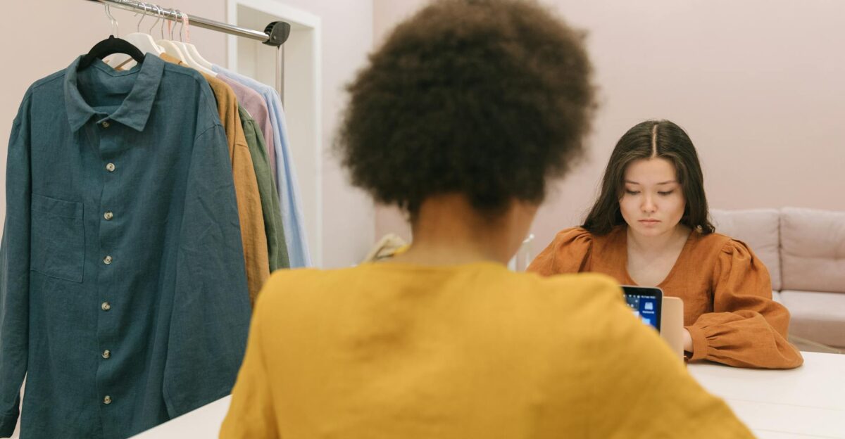 Two women working in a styled workspace with clothes rack and device focus