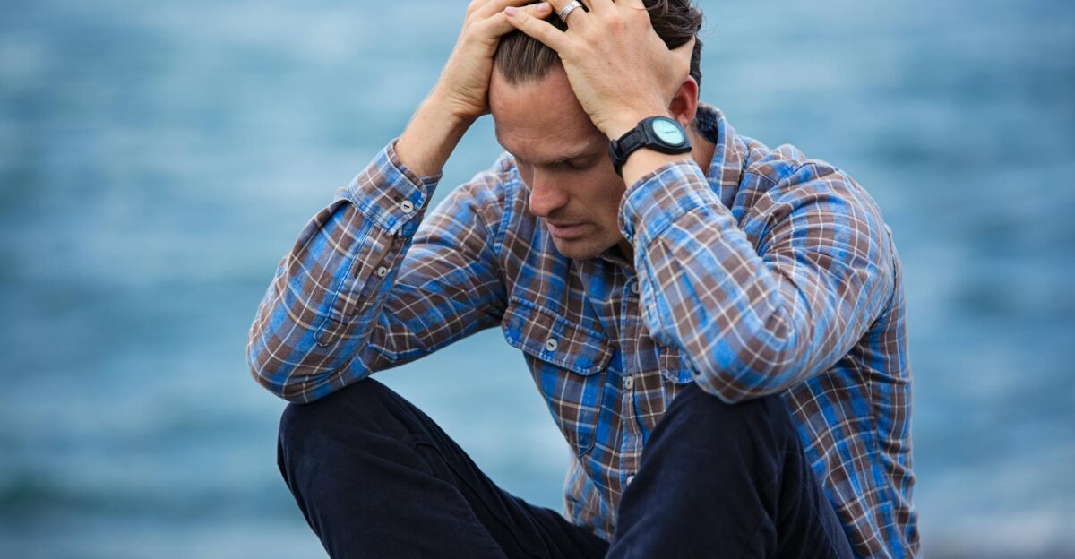 A man in a plaid shirt sits by the water looking distressed symbolizing stress