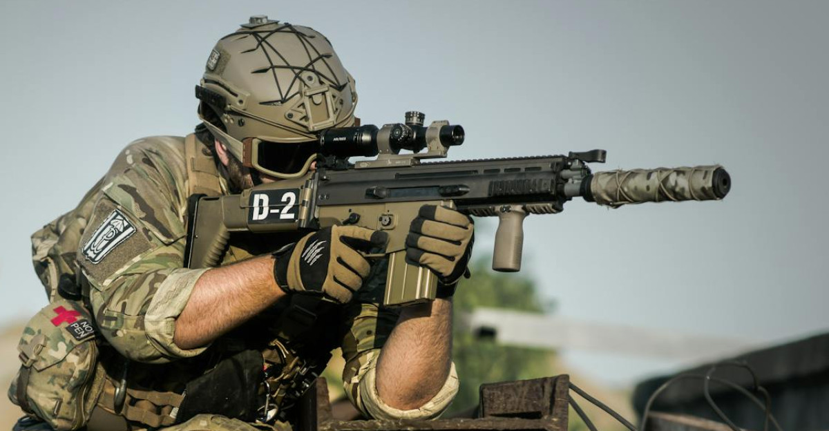 A soldier in full camouflage gear aiming a rifle while in a tactical stance outdoors