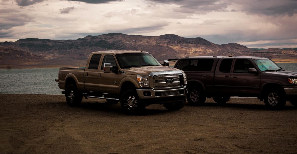 Pickup trucks parked by a serene mountain lake under a cloudy sky creating a rugged adventurous scene