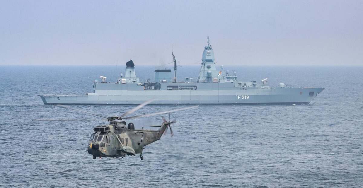 A military helicopter flies over the sea near a German warship F219