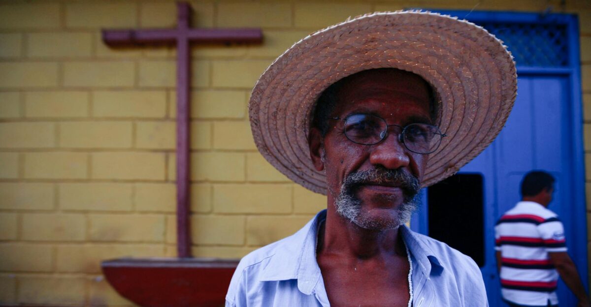 Close-up portrait of an elderly man wearing a straw hat standing near a church cross