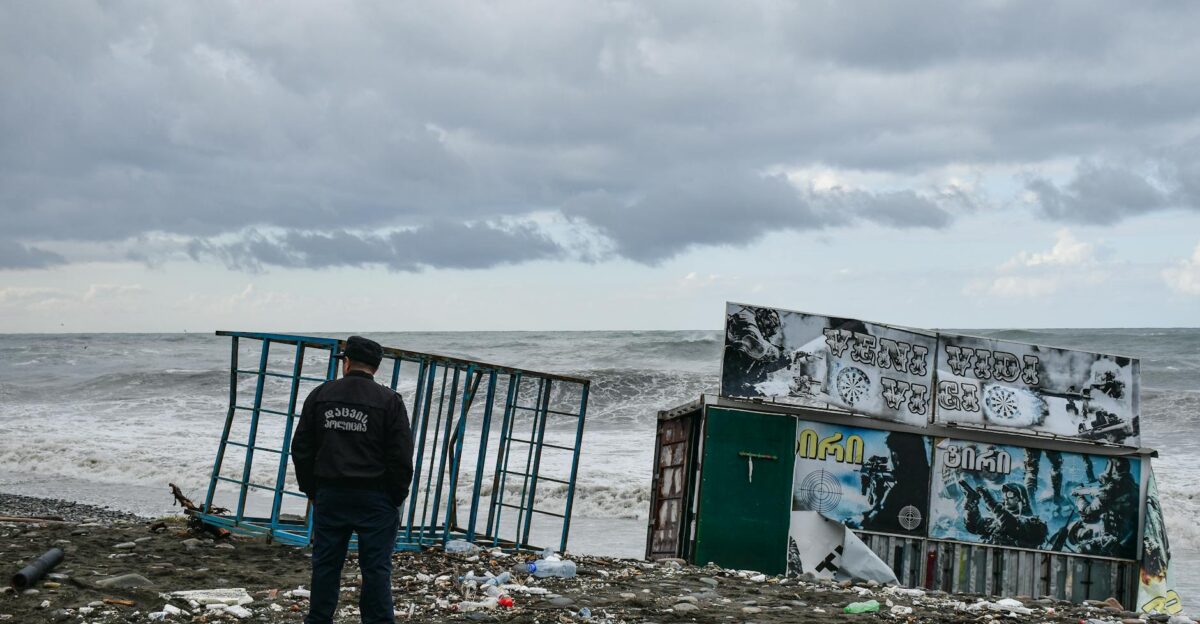 A man stands on a debris-filled beach observing a partially submerged structure