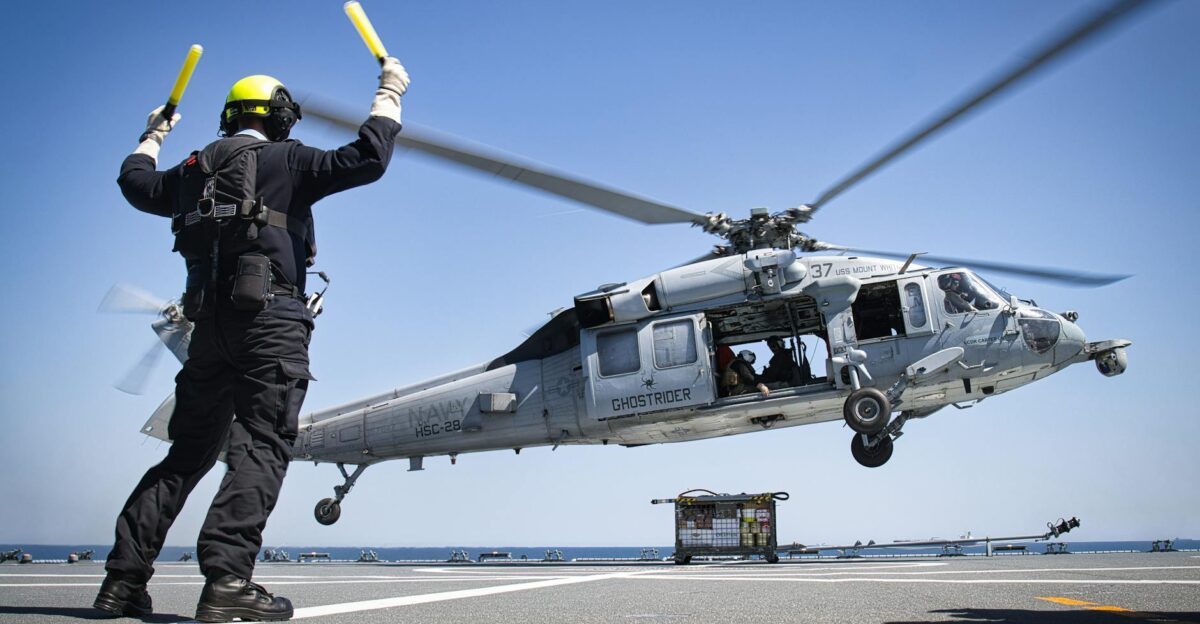 A military helicopter lands on an aircraft carrier guided by a deck crew member