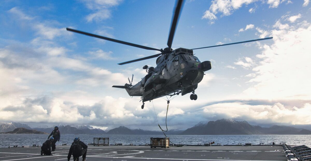 A military helicopter lands on a ship with mountains in the backdrop