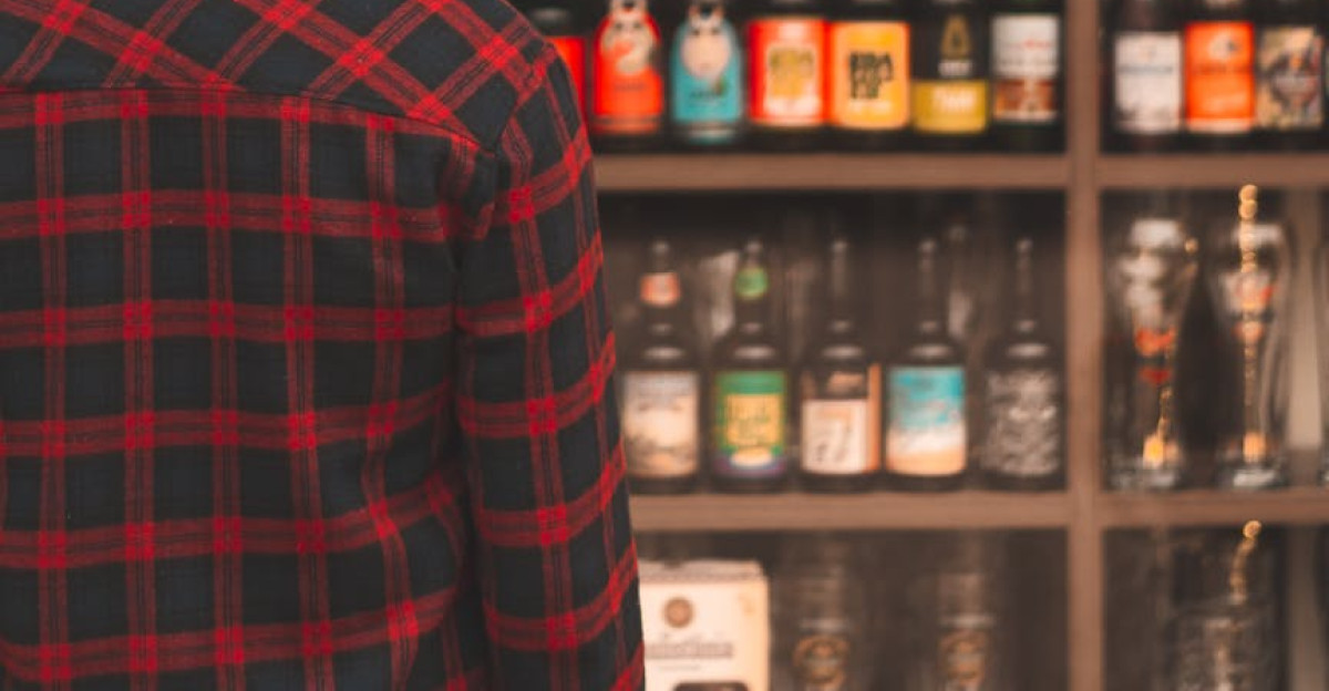 A man in a plaid shirt and hat examines a diverse beer shelf in a store