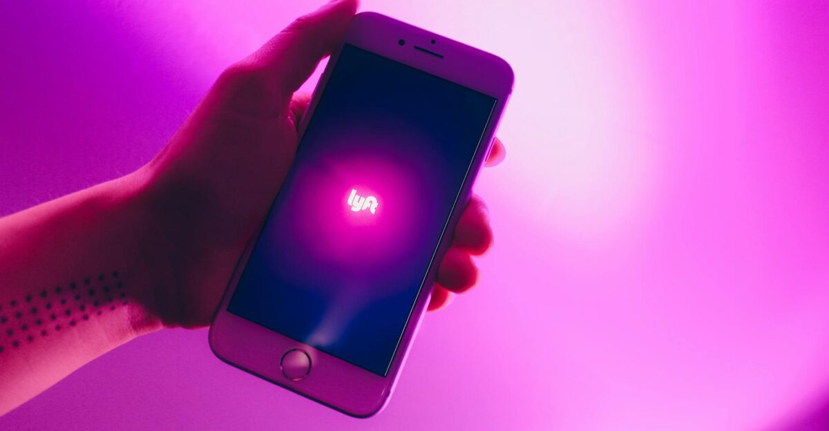 A hand holding a smartphone displaying the Lyft ridesharing app with a vivid pink background