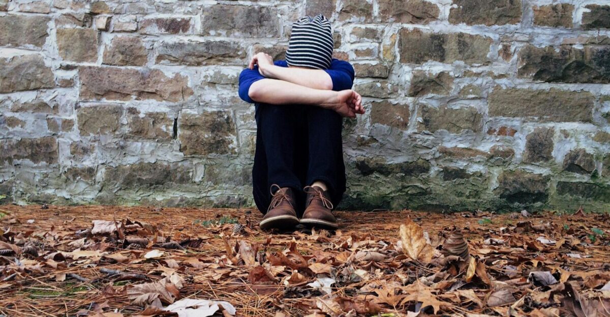 A solitary figure sits against a stone wall surrounded by fallen leaves expressing isolation