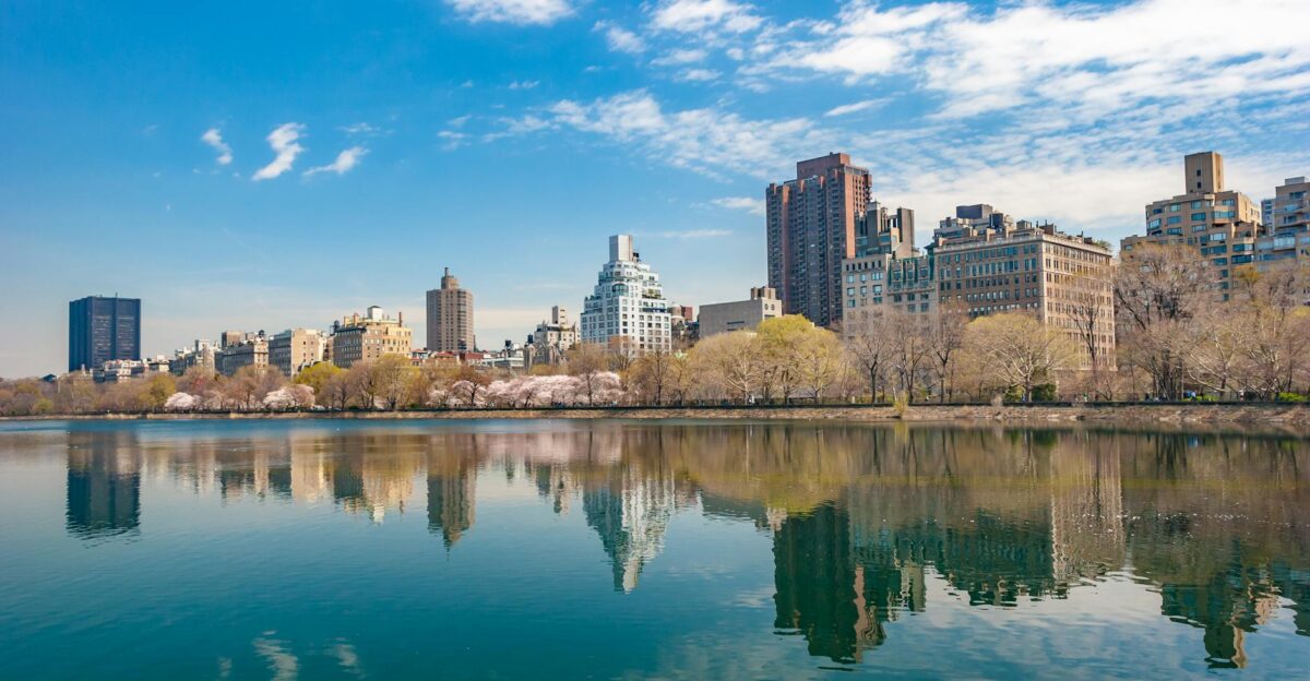 New York City skyline reflecting in Central Park s serene water under a vibrant blue sky