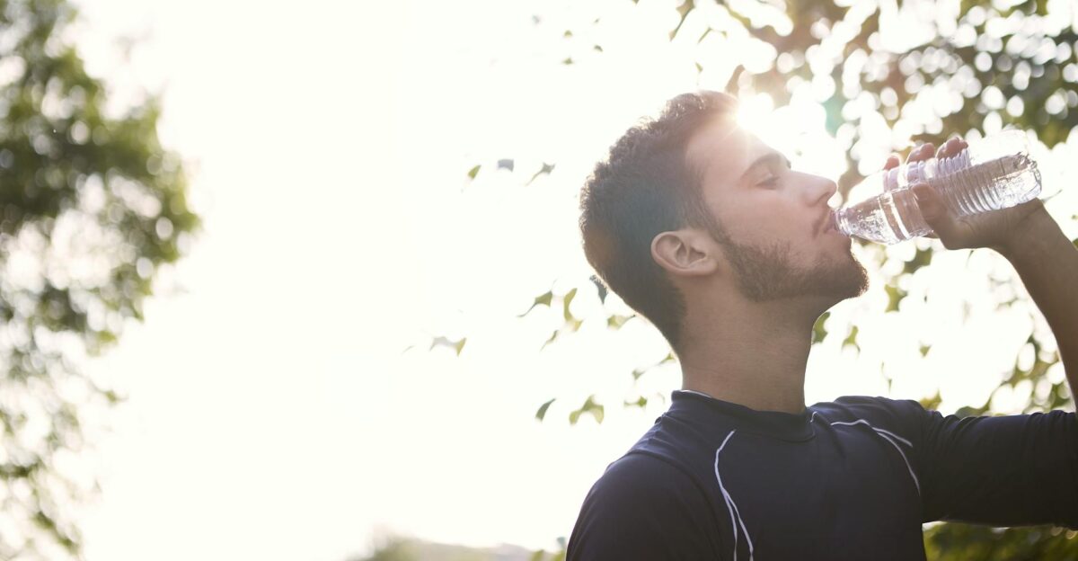 Side view of a young man drinking water from a plastic bottle outdoors in sunlight