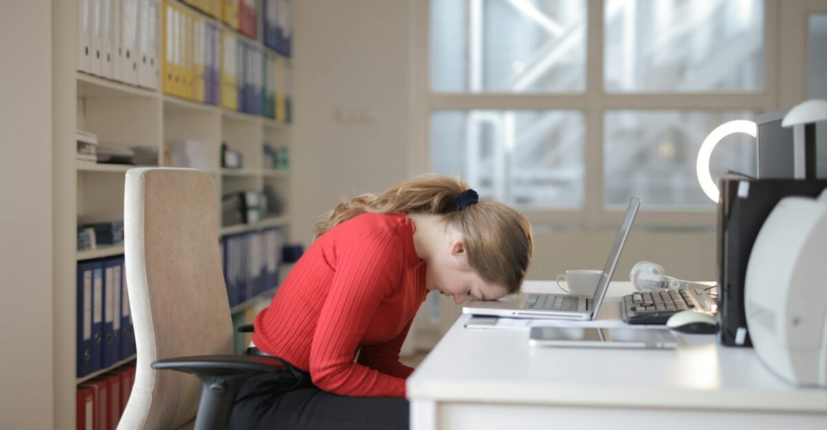Tired woman in red sweater naps on office desk beside laptop overwhelmed by remote work