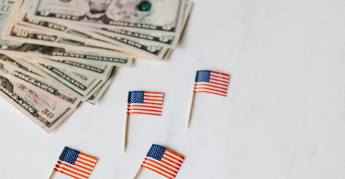 From above of flags of United States of America on toothpicks placed near bundle of American dollars of different nominal pars on white surface of table