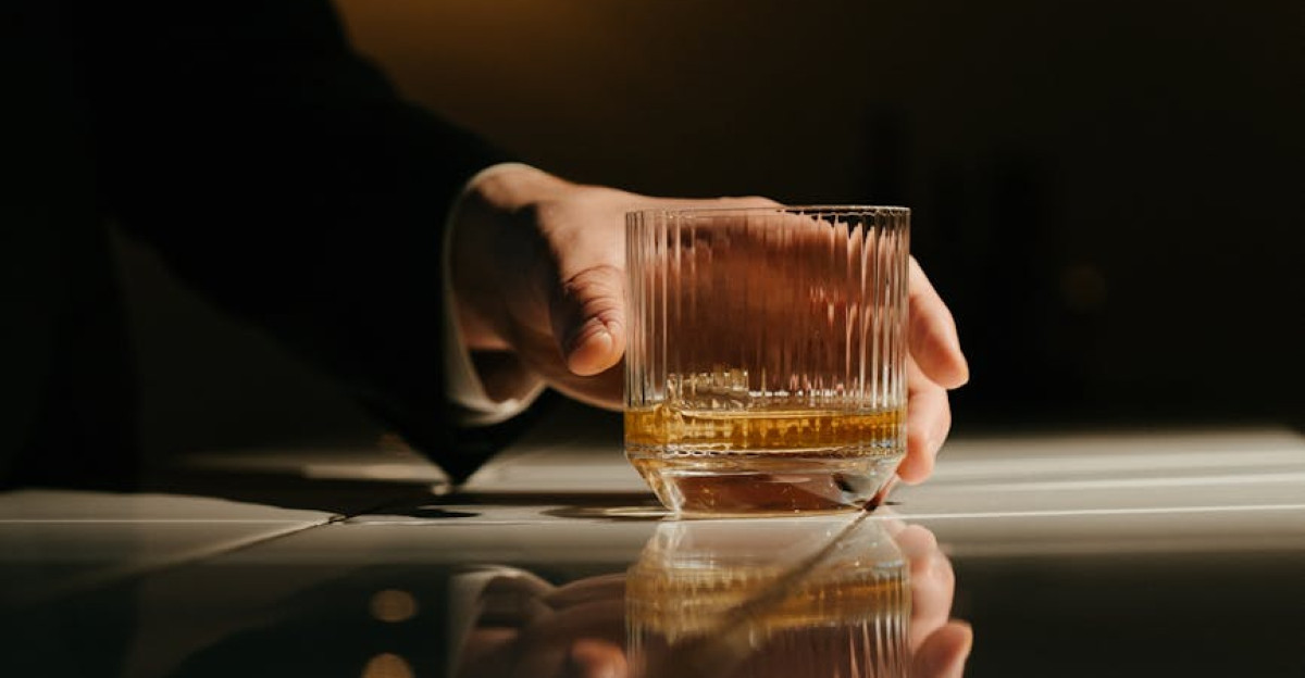 Close-up of a hand holding a whiskey glass with reflections on a bar counter in a dim light