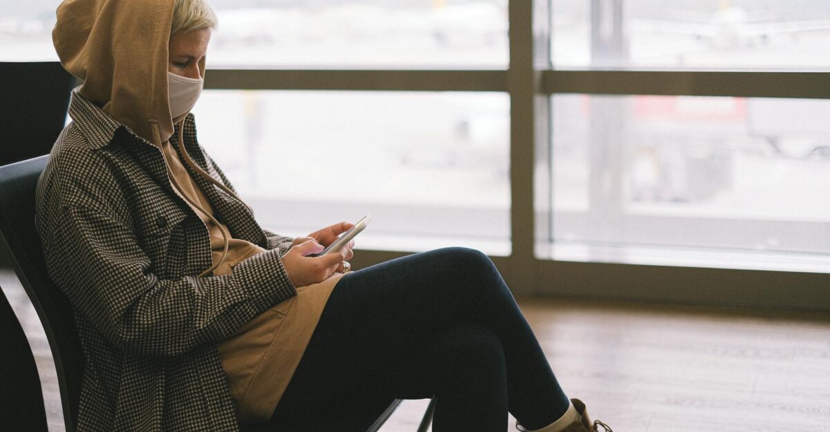 A woman in a hoodie and face mask uses a smartphone while waiting at an airport terminal