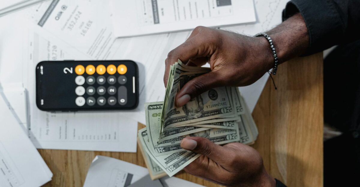 Person counting dollar bills over documents with a smartphone calculator on the desk