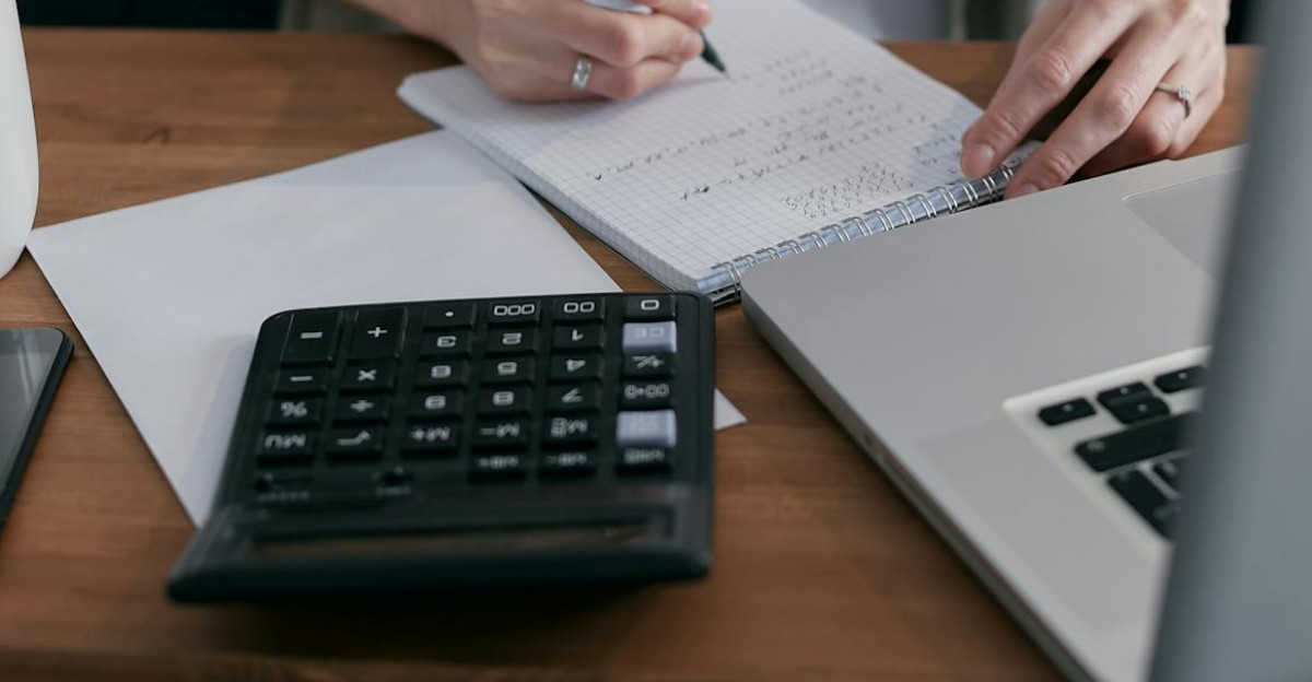 A woman writes financial calculations in a notebook using a calculator and laptop at a wooden desk