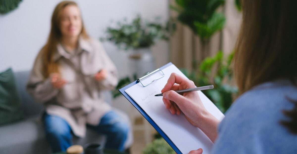 Unrecognizable professional female psychologist writing on clipboard while sitting against client on blurred background during psychotherapy session in light office
