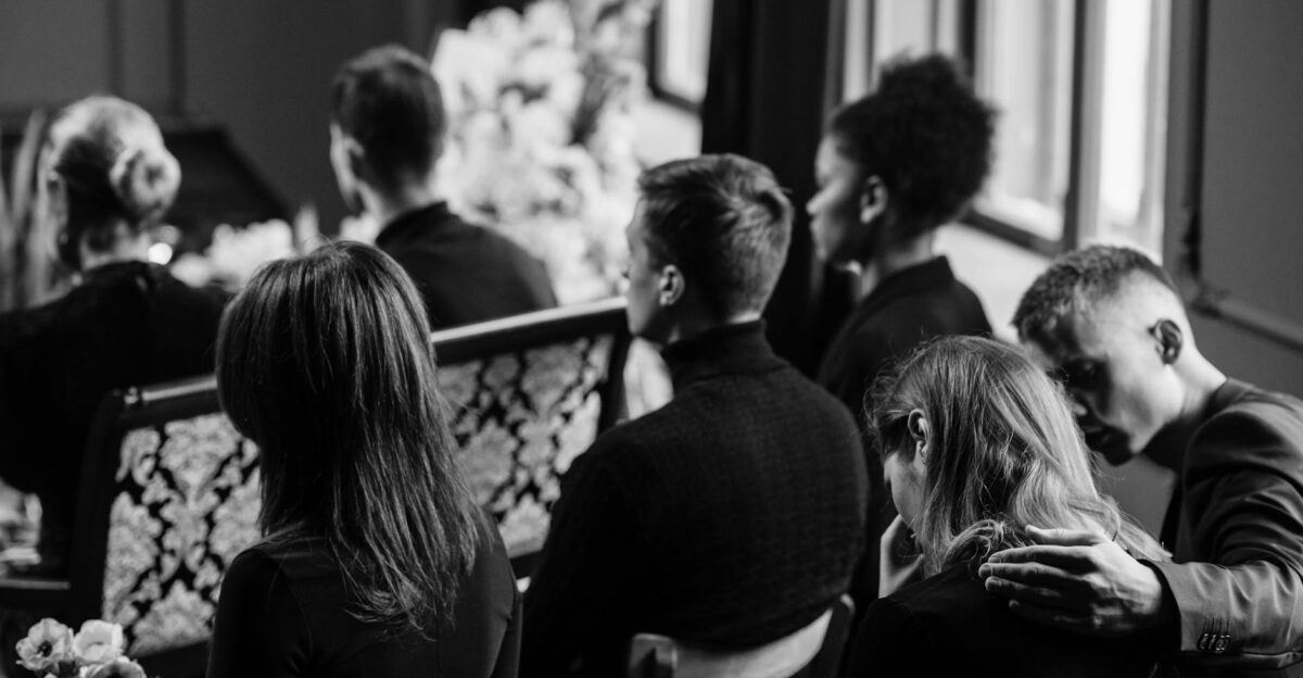 A somber grayscale image of mourners sitting indoors during a funeral conveying loss and sympathy