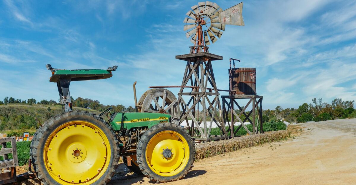 A green vintage tractor beside an old rustic windmill on a sunny rural farm