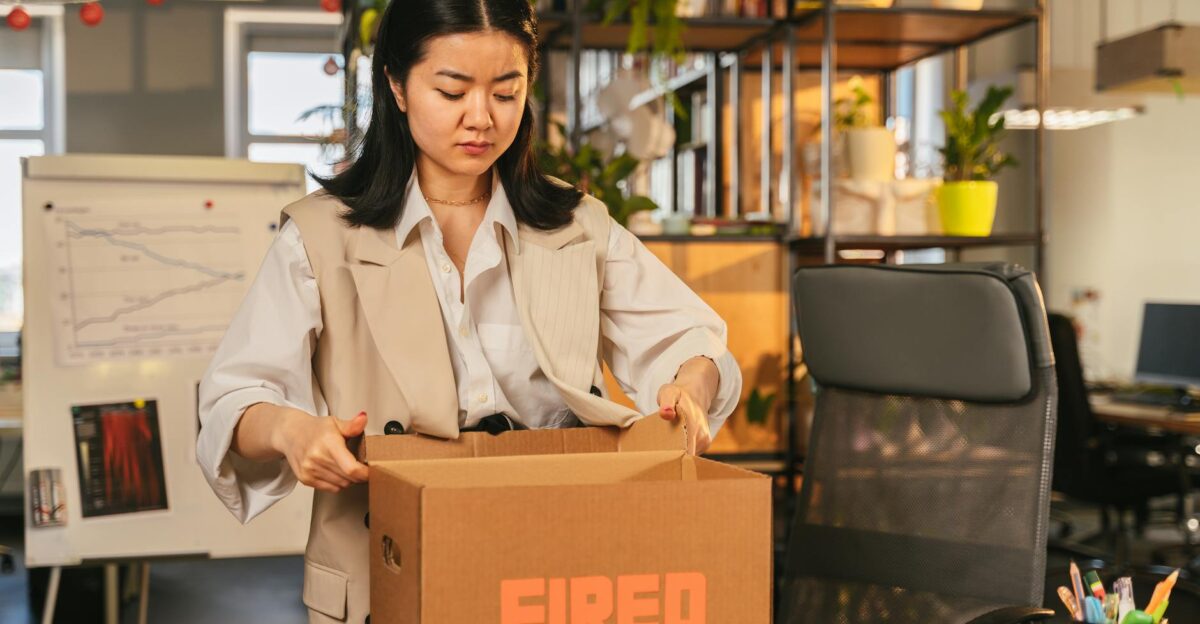 An Asian woman packs a box labeled FIRED symbolizing job loss in an office setting