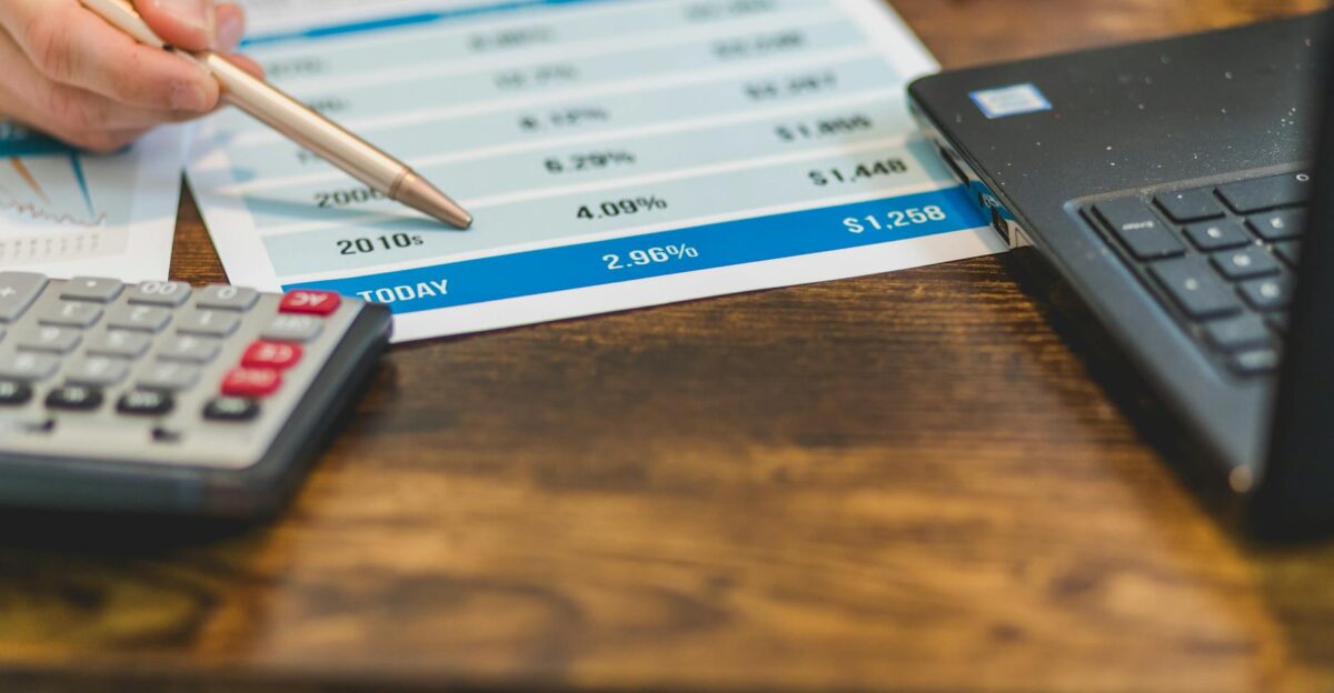 Close-up of a hand with pen analyzing financial rates on paper with a calculator and laptop nearby