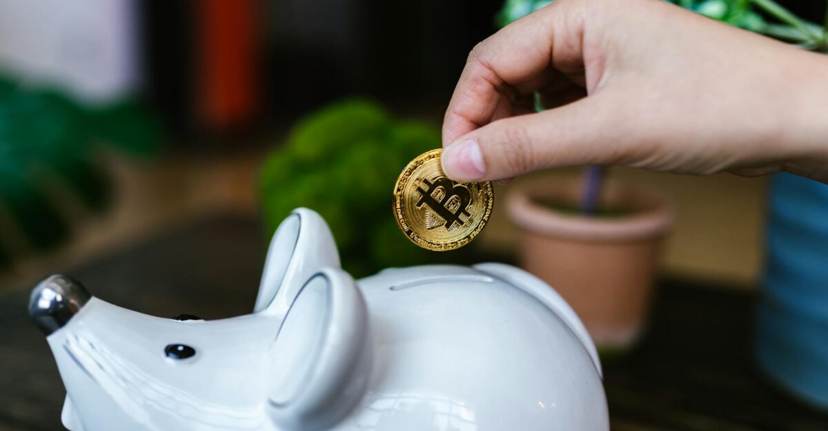 A hand placing a Bitcoin coin into a white piggy bank symbolizing savings