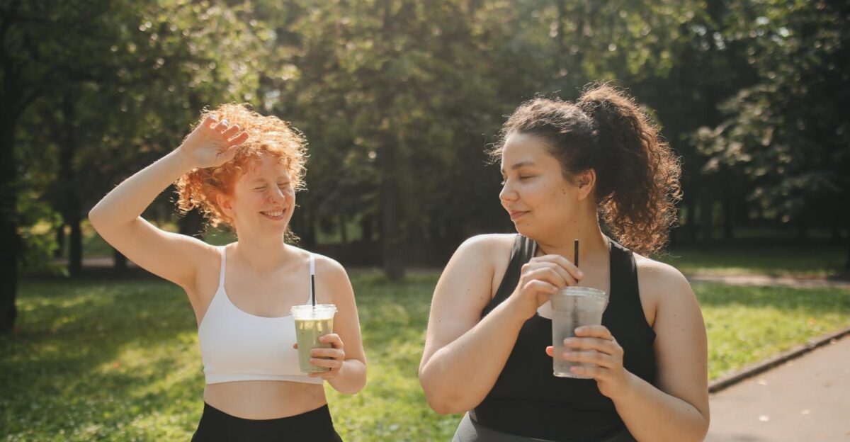 Two women in activewear enjoy refreshing drinks while relaxing outdoors in a sunny park