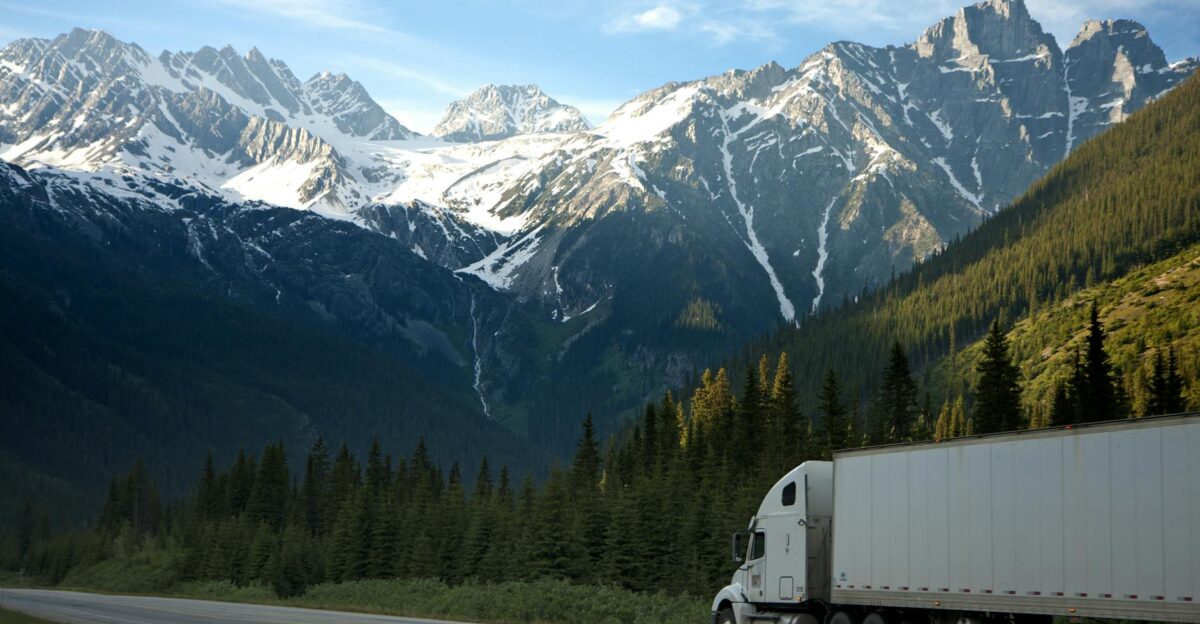 A semi-truck travels along a highway with snow-capped mountains in the background