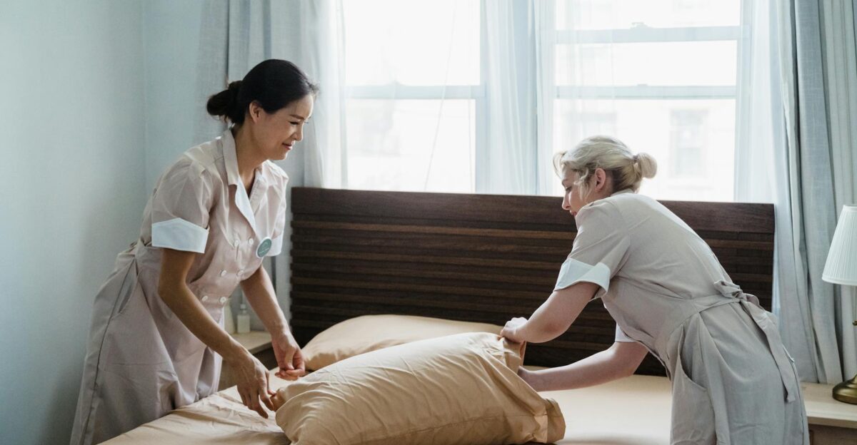 Two housekeepers in uniform collaborate while making a bed in a bright peaceful bedroom