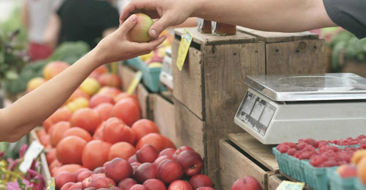 A customer exchanges an apple with a vendor at a vibrant farmers market