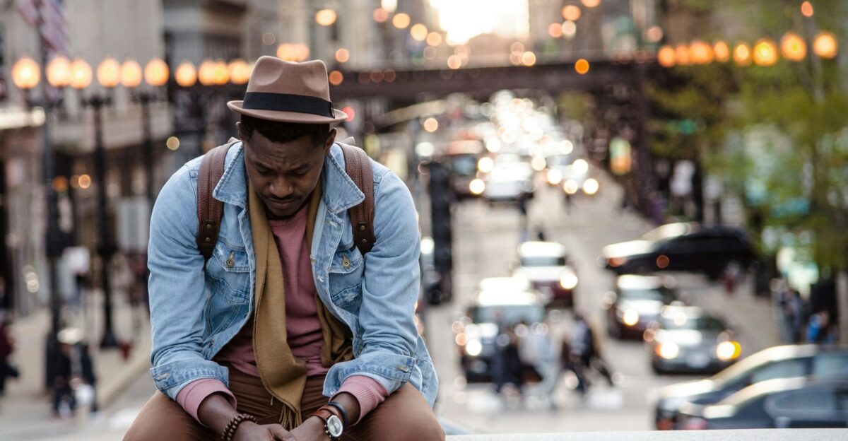 A man in a leather jacket looking down while sitting on a ledge in a city