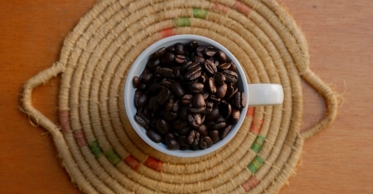 Coffee beans fill a white cup on a woven mat