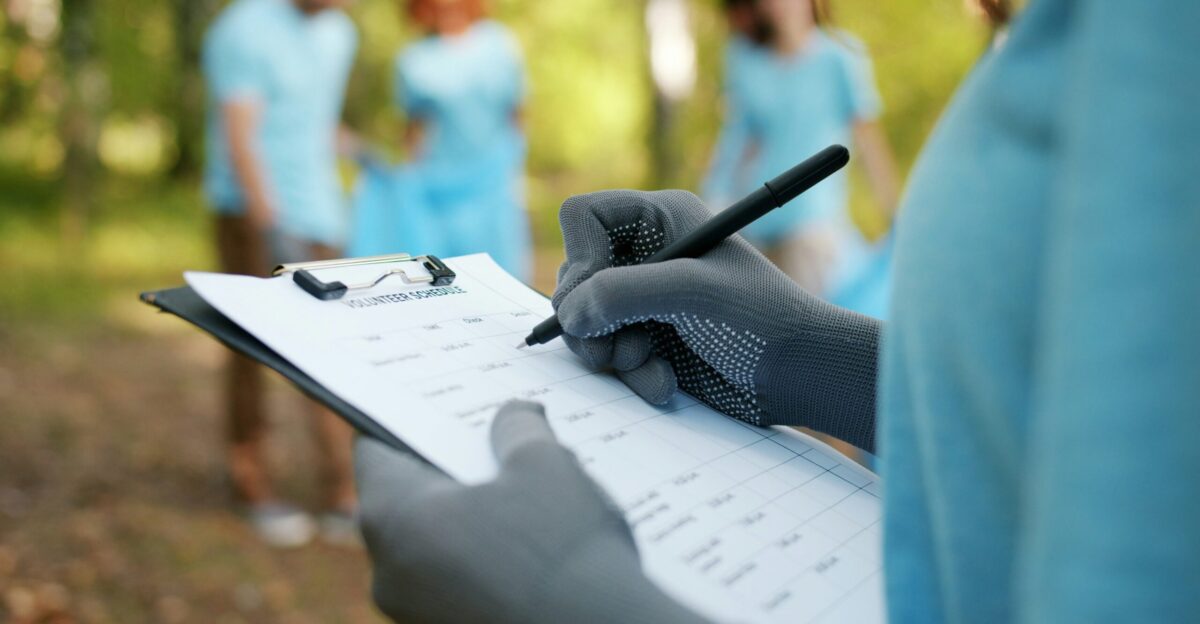 Person in gloves writing on clipboard with volunteers in background