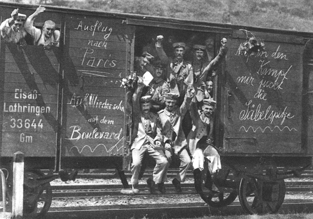 German soldiers some with flowers others waving or raising their clenched fists in a railroad car on the way to the front during early World War I 1914 Messages on the car spell out approximately Trip to Paris See you later on the Boulevard obscured by flowers the fight The obscured part most likely reads Auf in den Kampf which means Into battle my sword tip is itching