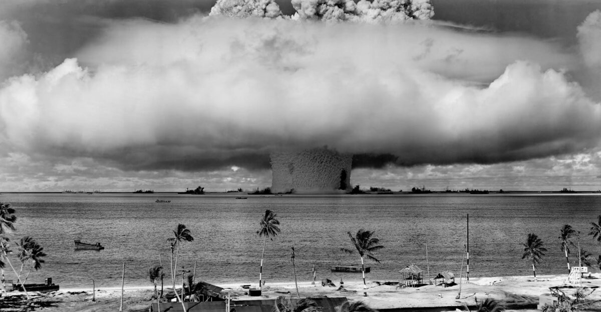 A black-and-white image of a nuclear explosion seen from a tropical beach with coconut trees