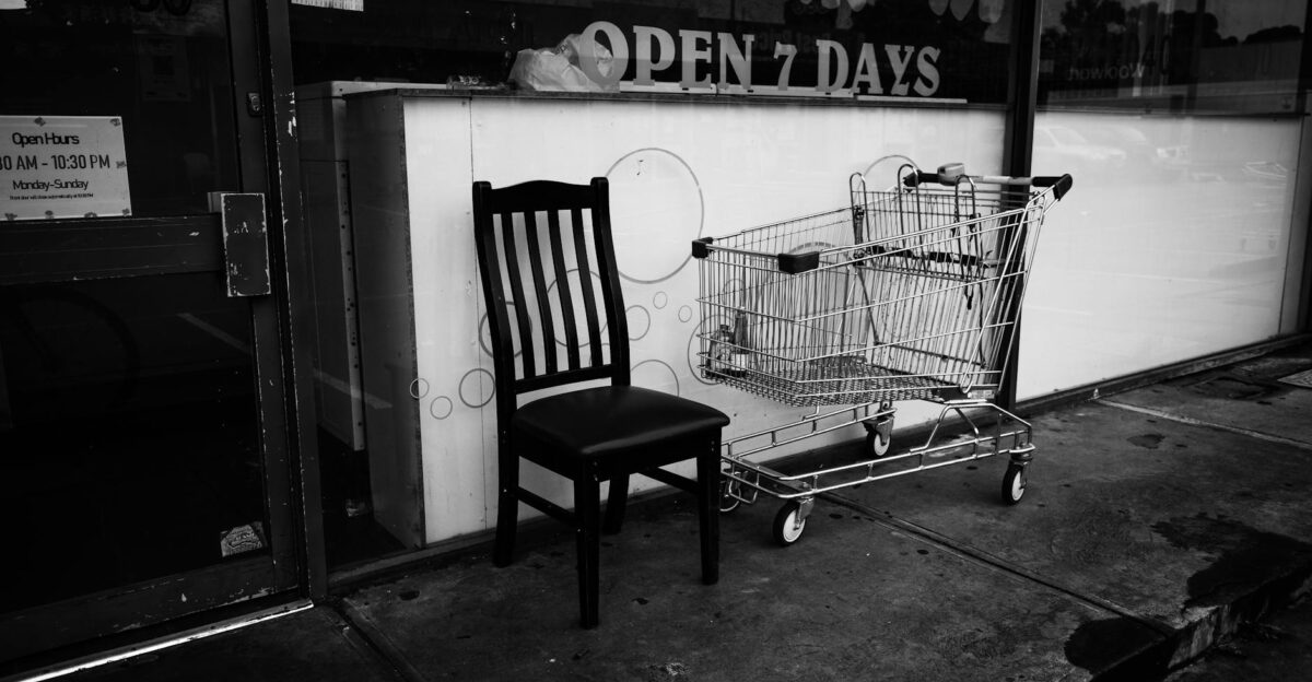 A black and white photo of a shopping cart and chair outside a store conveying an urban atmosphere