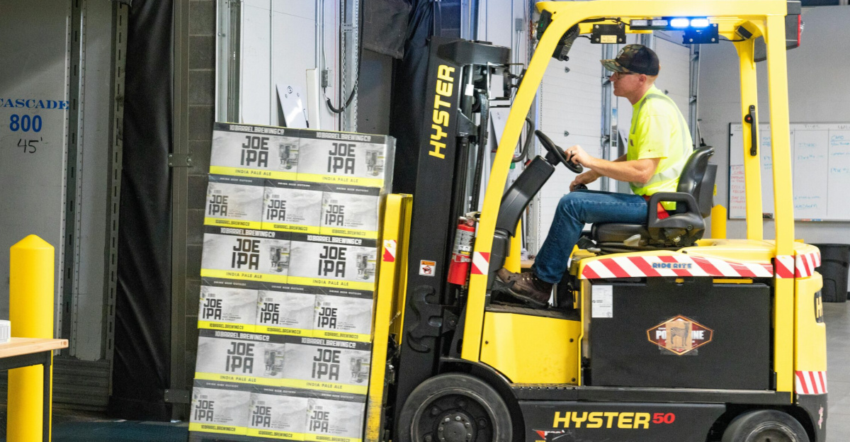 A worker drives a Hyster forklift moving Joe IPA boxes in a warehouse.