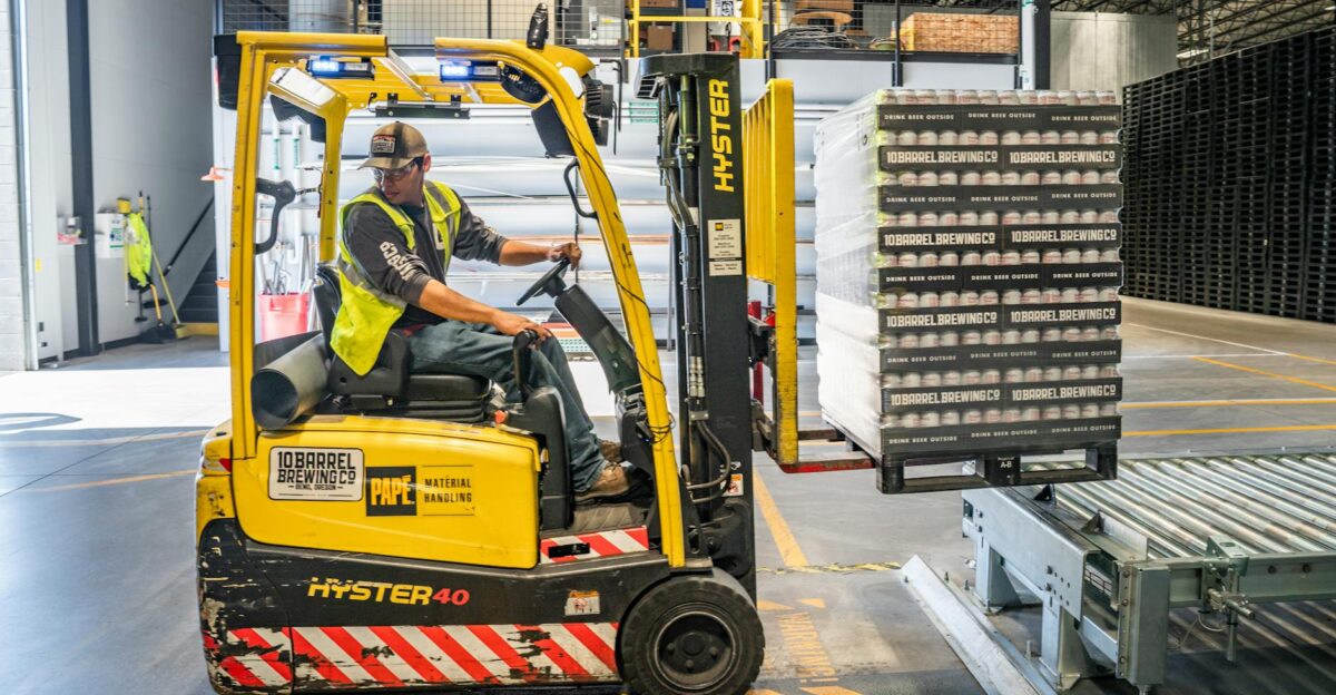 A warehouse worker maneuvers a forklift to transport crates for brewing company storage