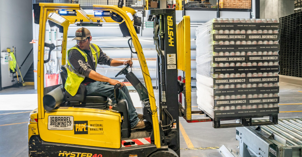 A warehouse worker maneuvers a forklift to transport crates for brewing company storage.
