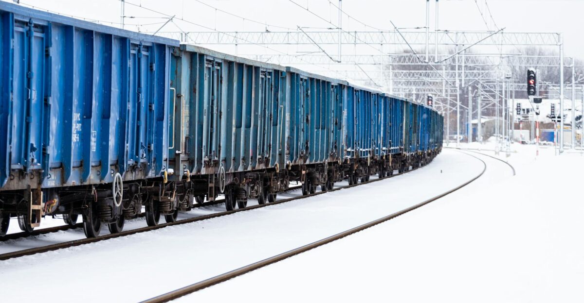 A blue cargo train travels on a snowy railway track in winter with overhead power lines
