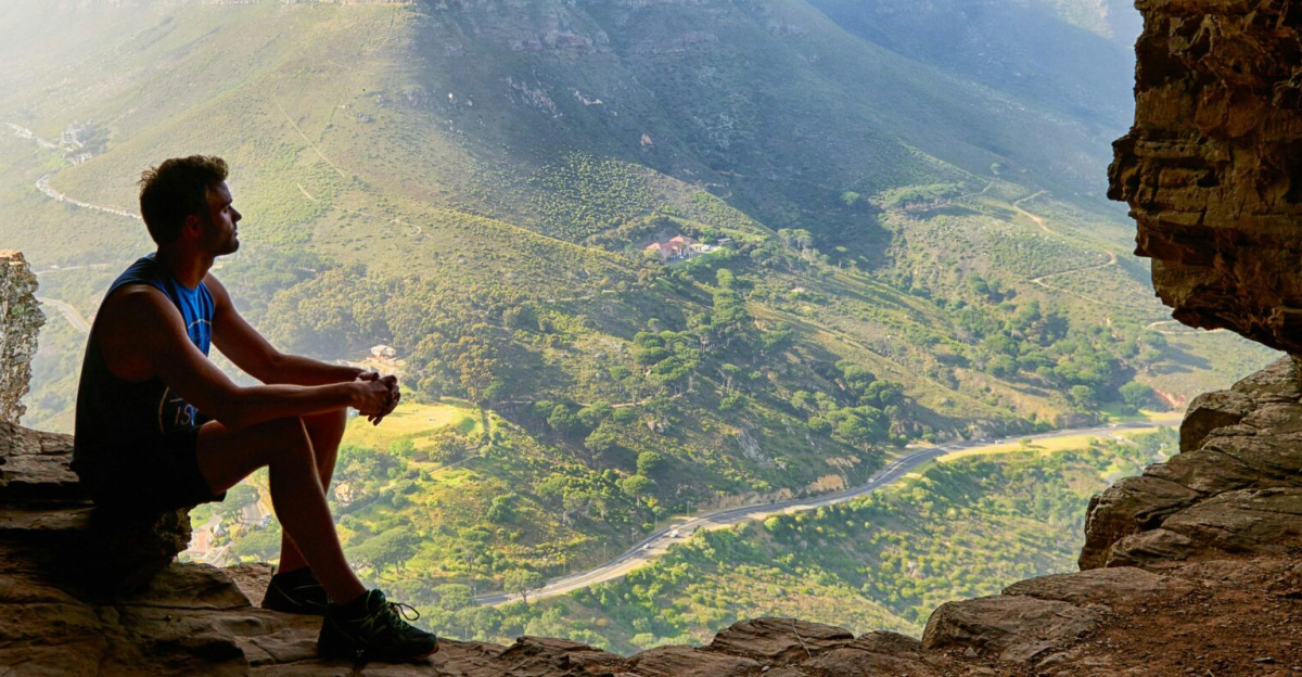 A man sitting in a cave overlooking a majestic mountain landscape under daylight.