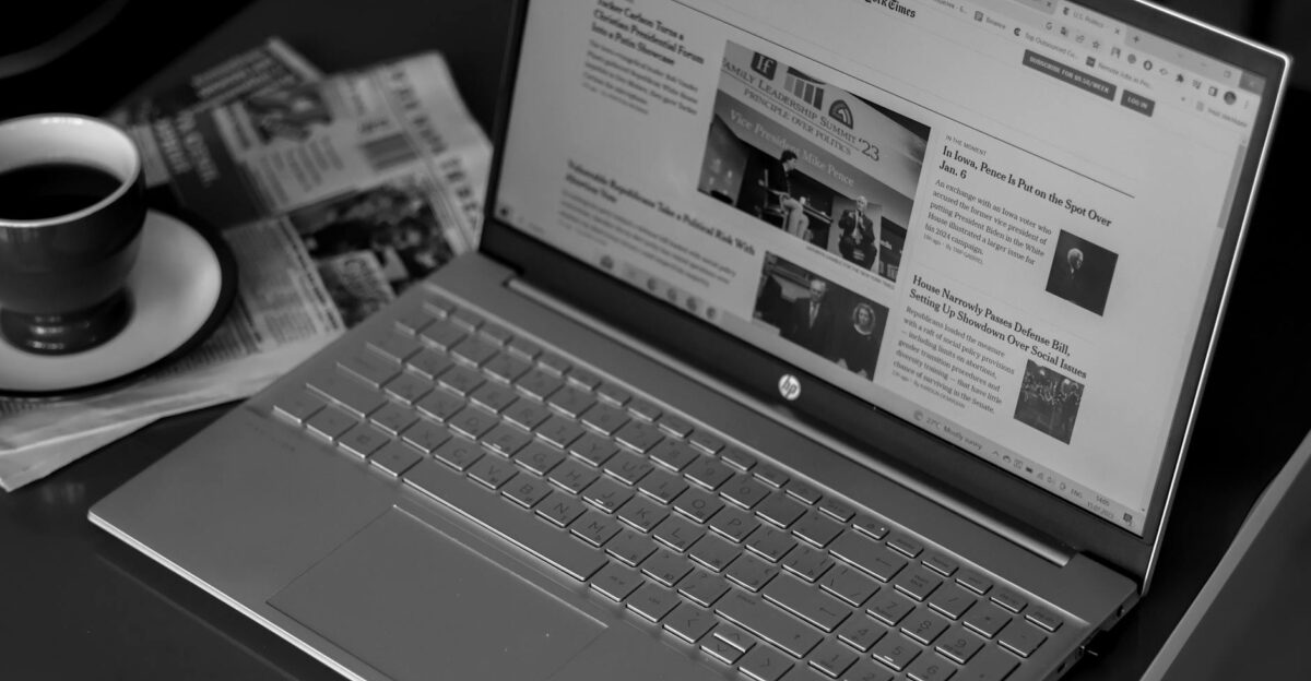 Black and white image of a laptop displaying news articles accompanied by a cup of coffee and newspapers