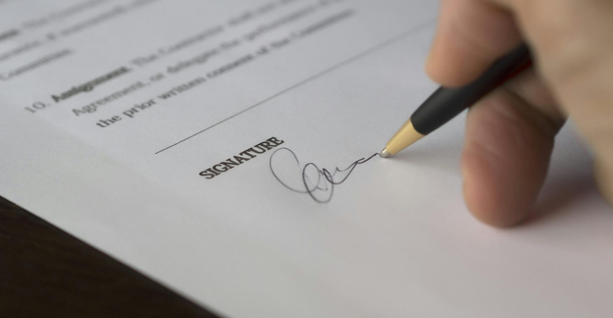 A hand signs a formal contract with a pen on a wooden desk.